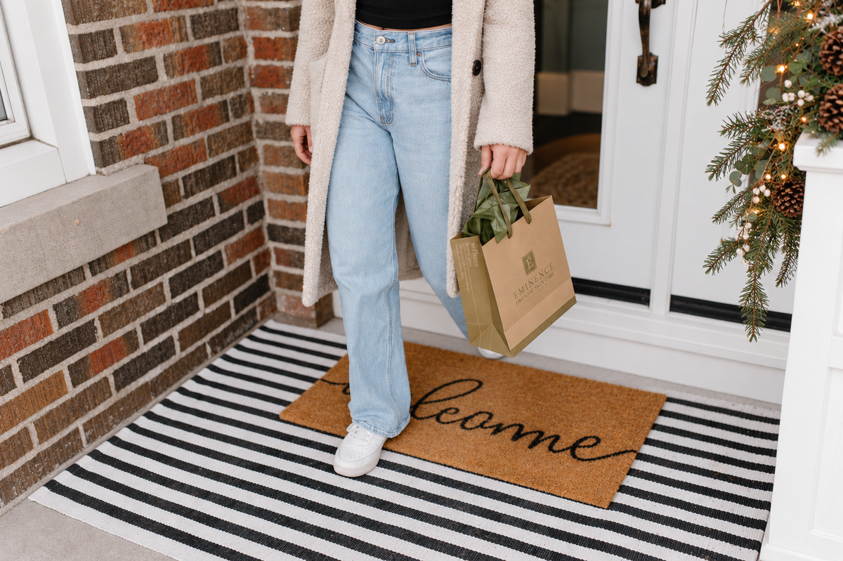 Shopper in a shearling coat leaving Just Us Boutique with an Eminence gift bag, standing on a welcome mat by a holiday garland